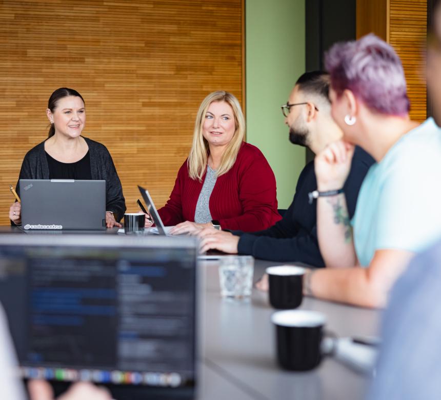 Mehrere Personen sitzen an Besprechungstischen zusammen, mit Notebooks vor sich, und sind in intensiver Diskussion