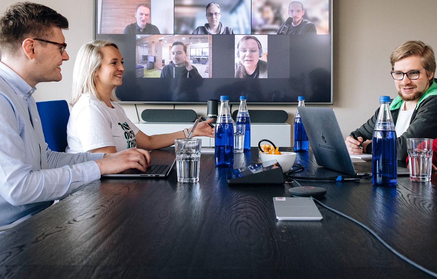 Project meeting: three people in a meeting room, 5 others present via video conference on the screen on the wall