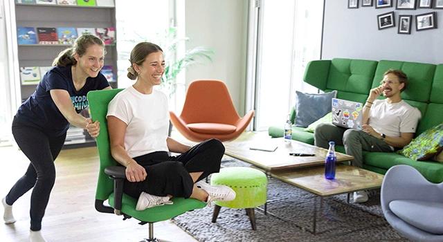 Lachende Person wird von einer Weiteren auf Stuhl durch die Gegend gerollt, im Hintergrund noch eine Person, die lacht