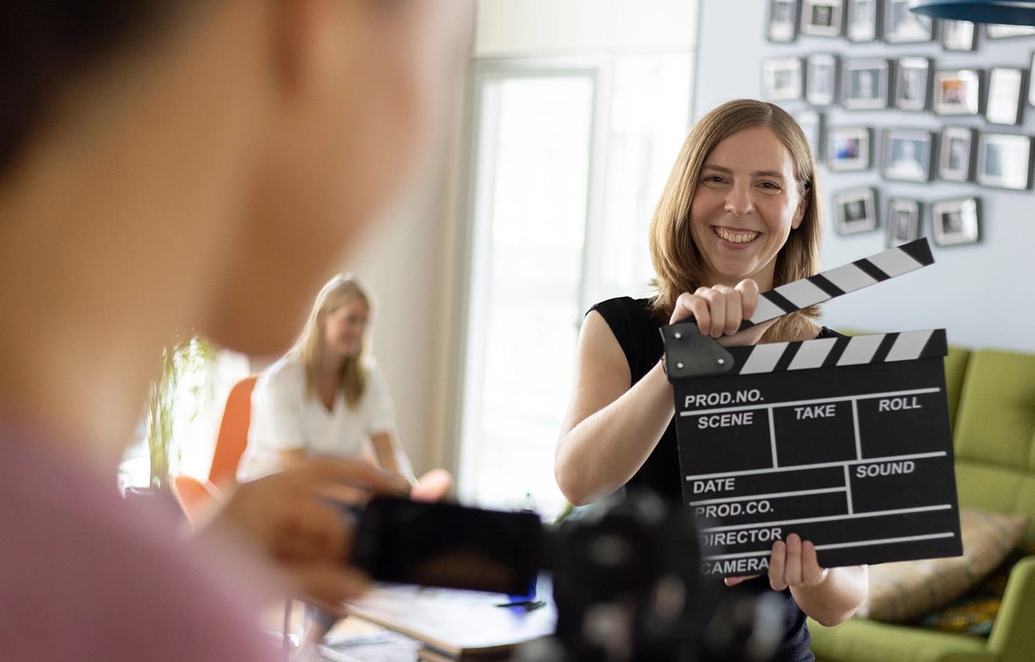 Eine Person im Fokus, lachend mit einer Filmklappe in der Hand. Eine weitere unscharf zu erkennende Person filmt diese.