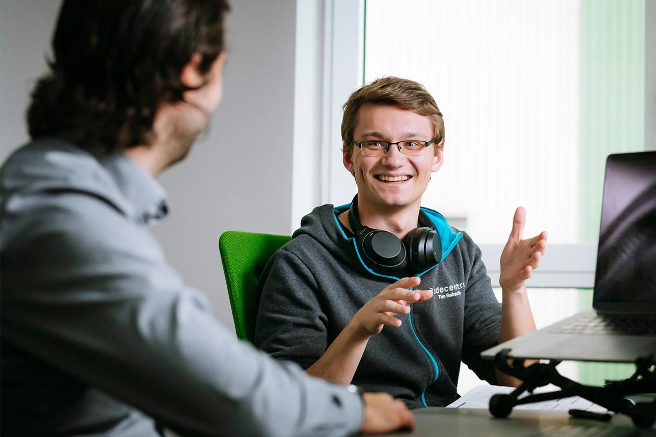 Zwei Menschen sitzen zusammen an einem Arbeitsplatz und unterhalten sich gut gelaunt