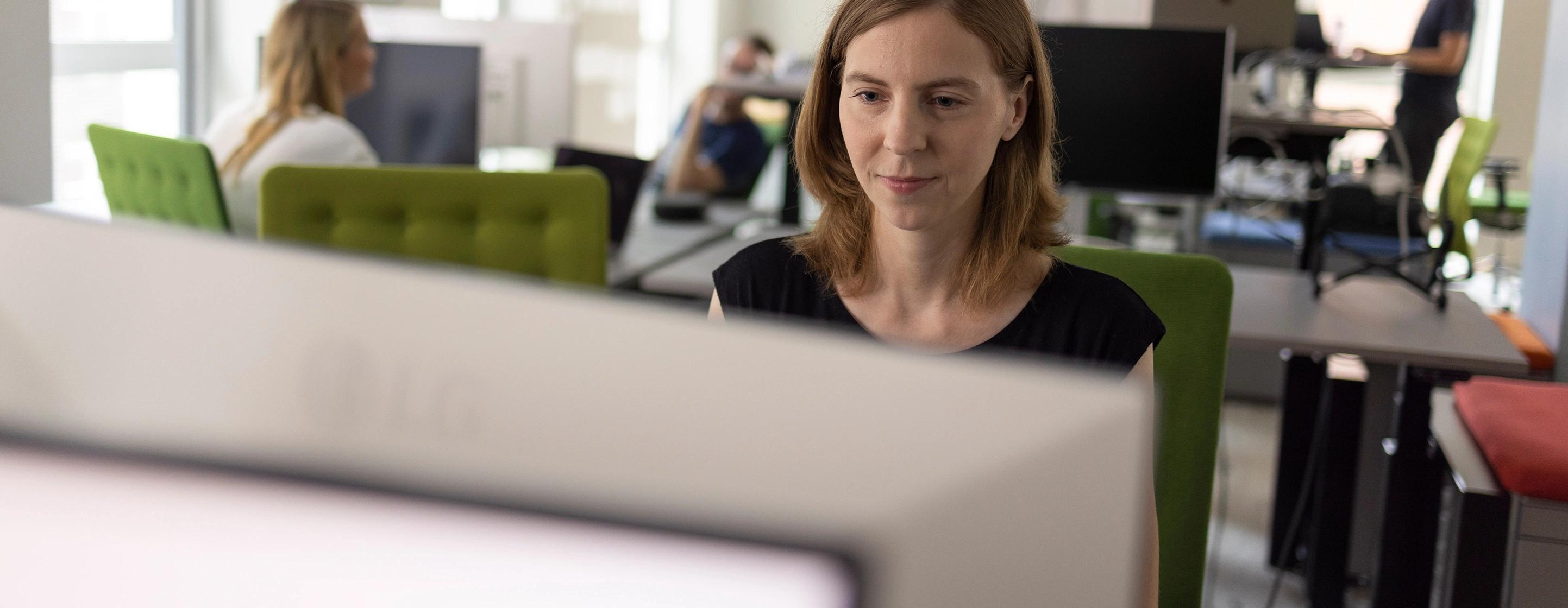 Eine Frau sitzt an einem Computer und arbeitet fokussiert, im Hintergrund sind verschwommen noch weitere Personen zu sehen