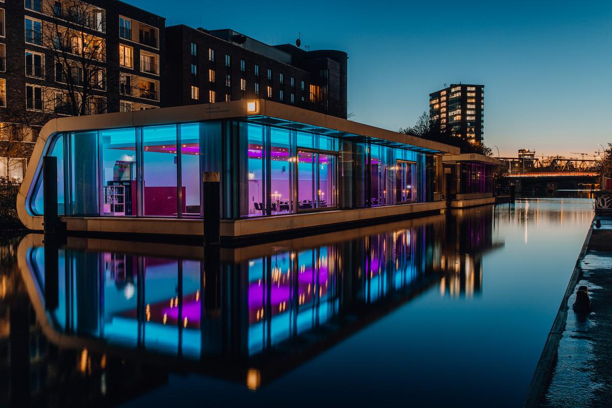 Ein von innen bunt beleuchtetes, flaches Gebäude in der Dunkelheit, was auf einem Fluss in der Innenstadt steht.