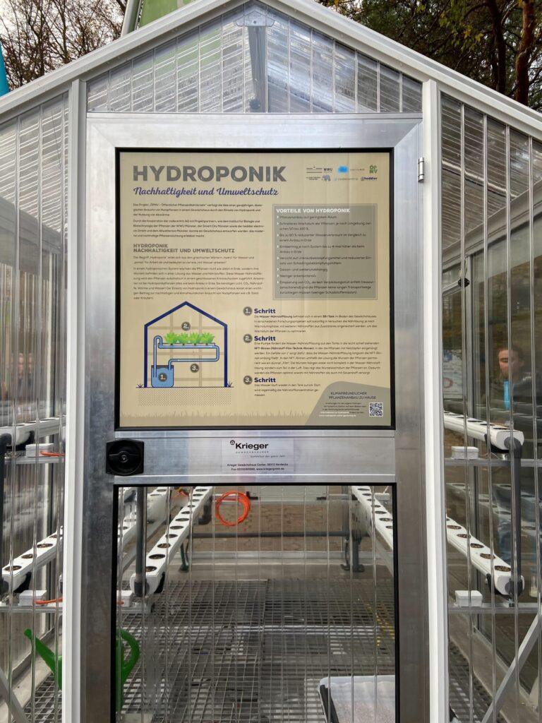 Ein leeres Gewächshaus aufgenommen von außen, mit einem Schild an der Tür wo erklärt wird wie das Gewächshaus funktioniert.