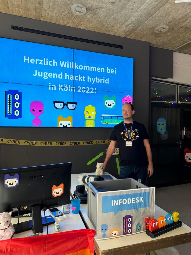 Martin Wildau stehet an einem Infodesk mit vielen kleinen bunten Figuren, beim Jugend hackt hybrid 2022.