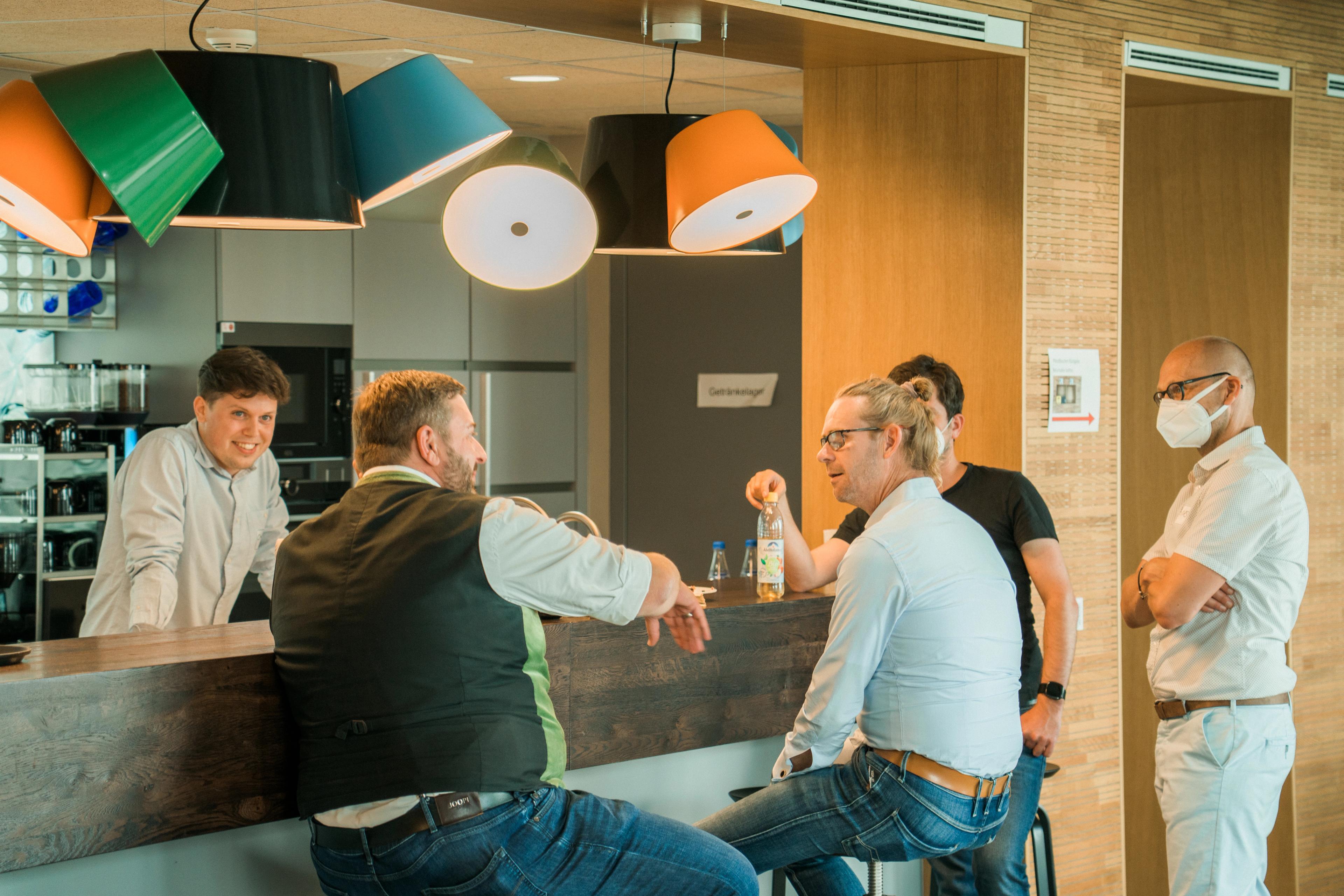5 Personen an Theke einer Cafeteria, 2 Sitzend und 3 stehend, Unterhalten sich.
