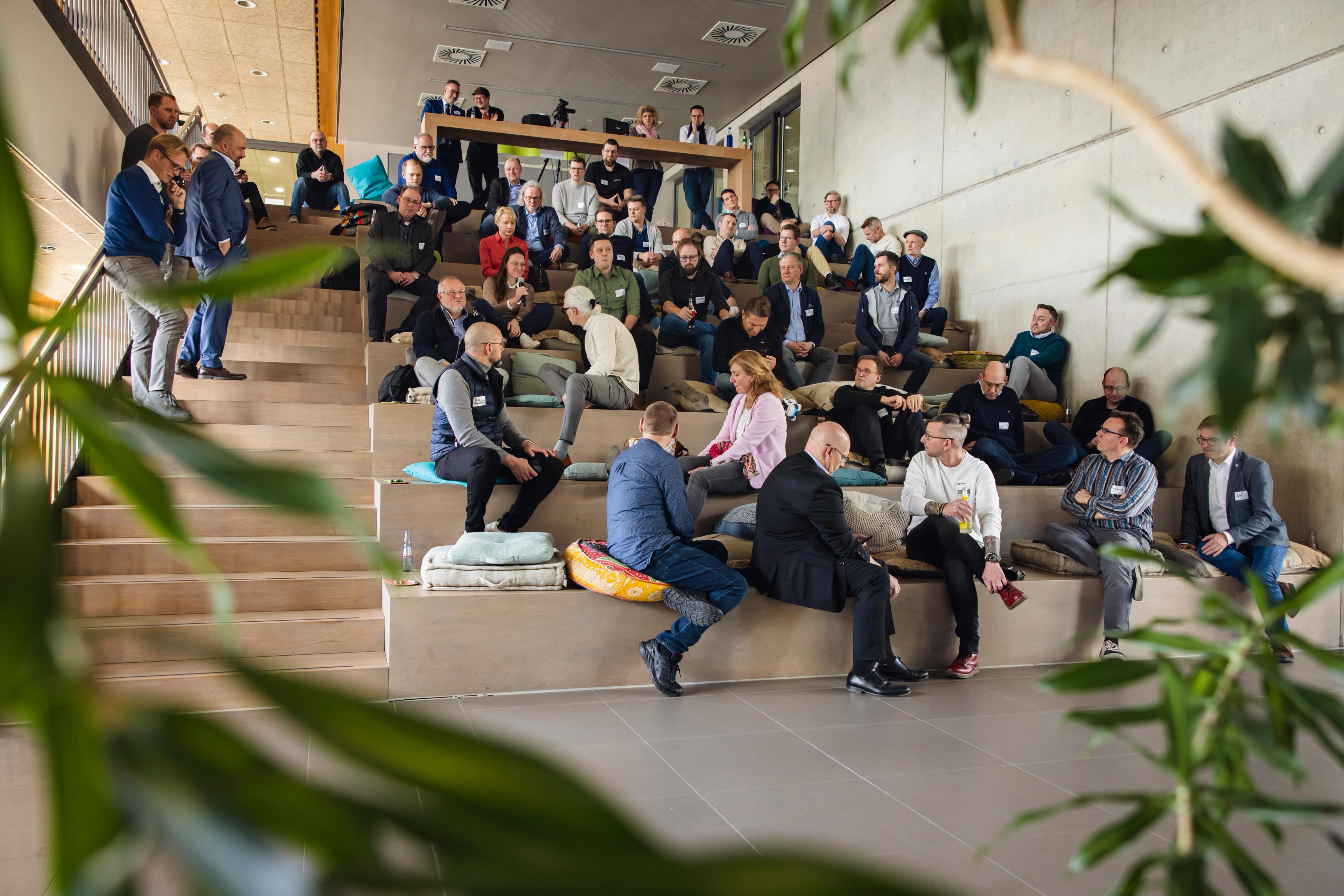 Viele Personen in einem kleinen Auditorium gucken einen Vortrag, Frau mit Mikrofon zwischen den Personen spricht.