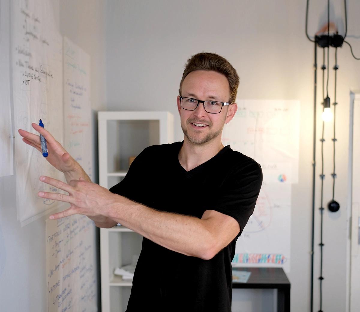 Eine Person vor einem beschrifteten Whiteboard, mit Stift in der Hand, schaut in die Kamera.