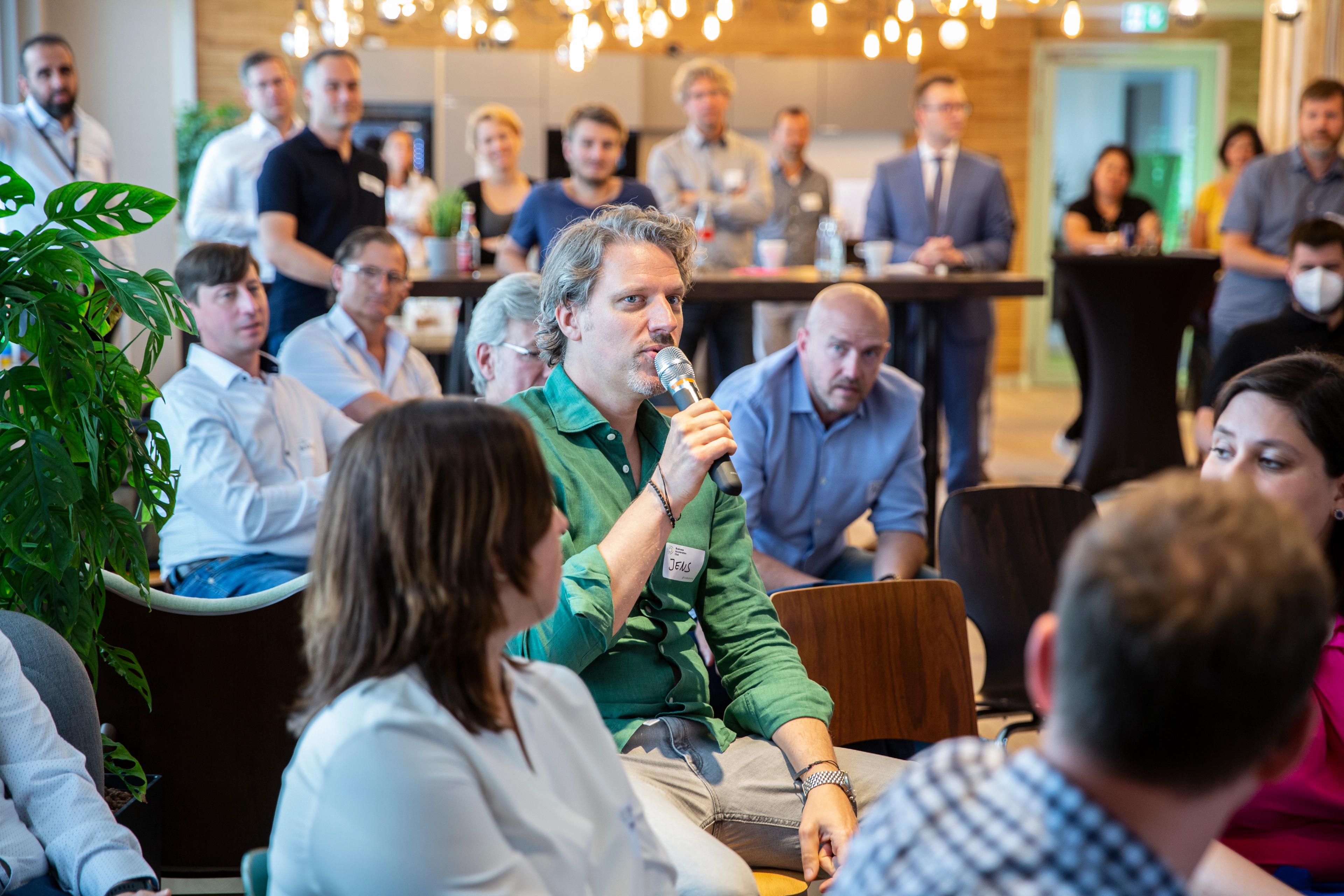 Eine Person des Business Acceleration Club, die inmitten vieler sitzender und stehender Personen mit einem Mikrofon Spricht.