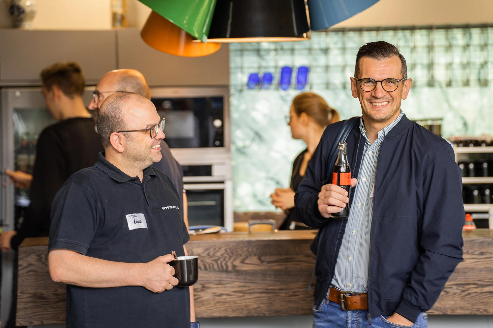 2 Personen, die mit Softgetränk und Cafe vor der Küche einer Cafeteria stehen und lachen, 3 weitere Personen im Hintergrund.